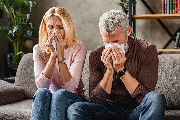 Middle aged couple sitting on sofa blowing their nose , flu concept with tissues.