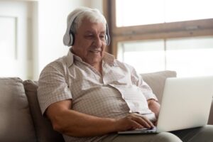 A smiling old man wearing headphones and using a laptop at home to take an online hearing test.