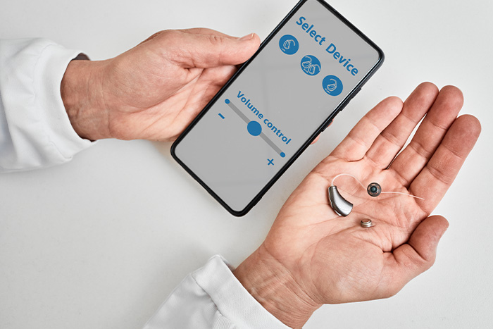 Audiologist showing smartphone app for adjusting hearing aid holding smartphone in one hand and BTE hearing aid in other.